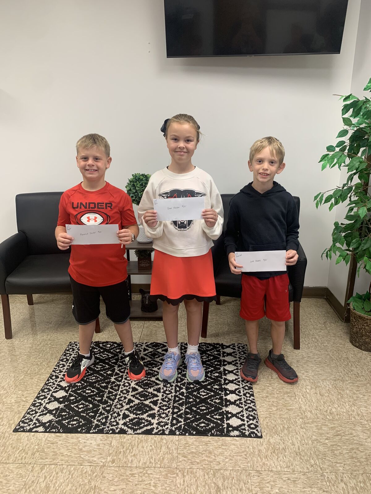 Three children stand proudly indoors, holding white cards with handwritten text while posing for the camera.
