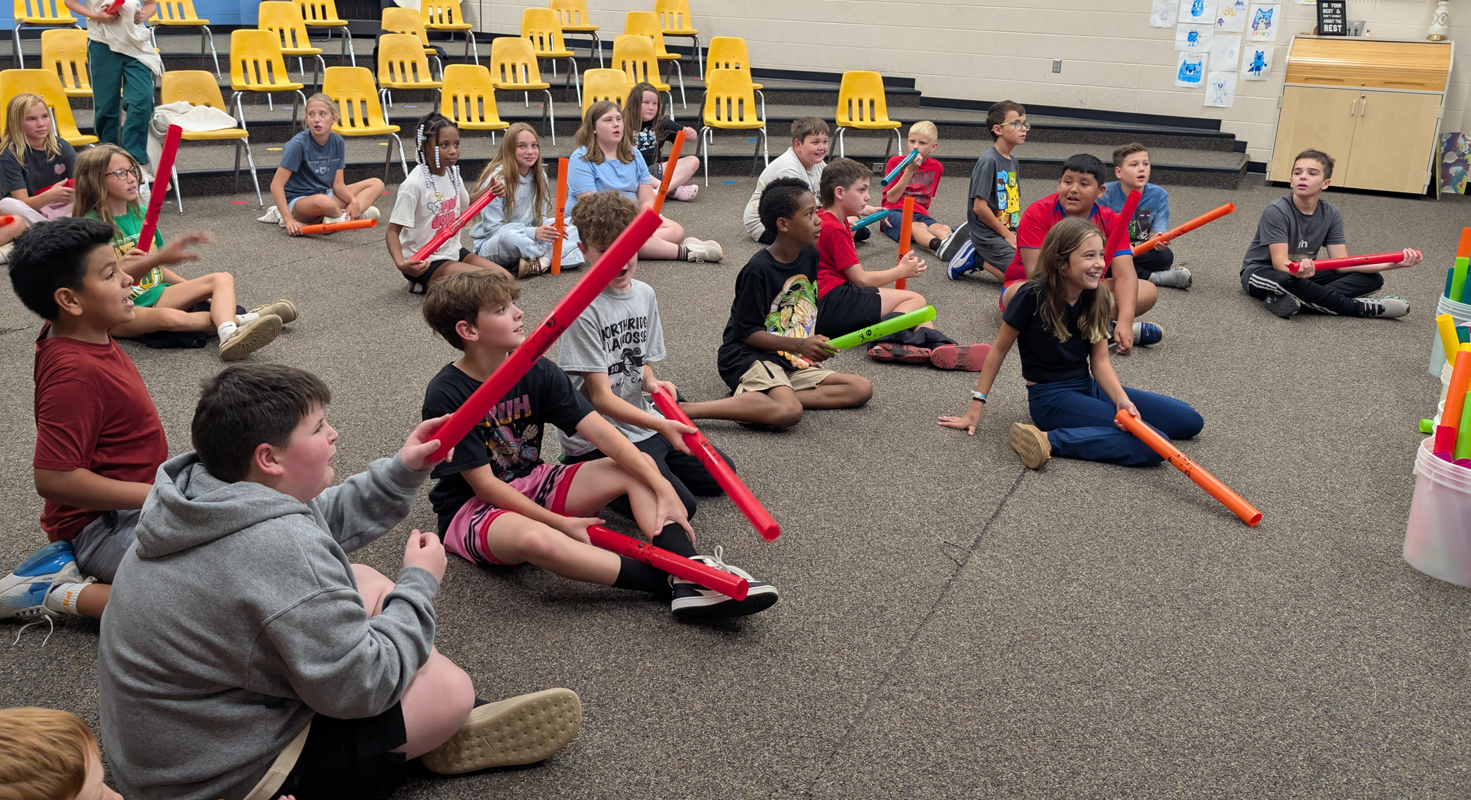 Children sit on the floor holding colorful foam tubes, engaged in an activity.