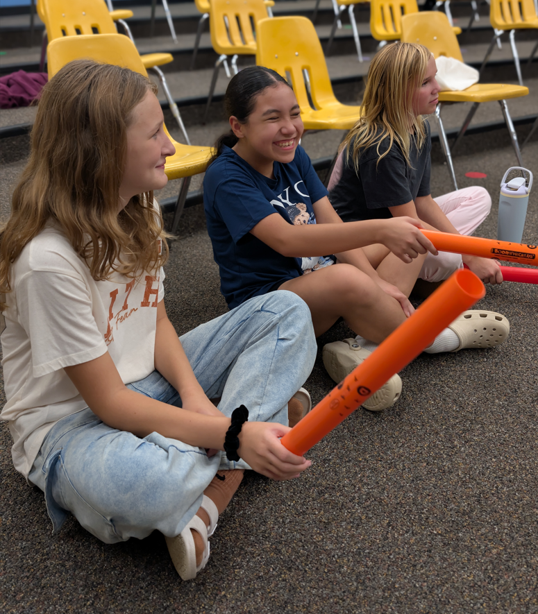 Three girls sit on the ground in front of yellow bleacher seats, holding orange noisemakers.
