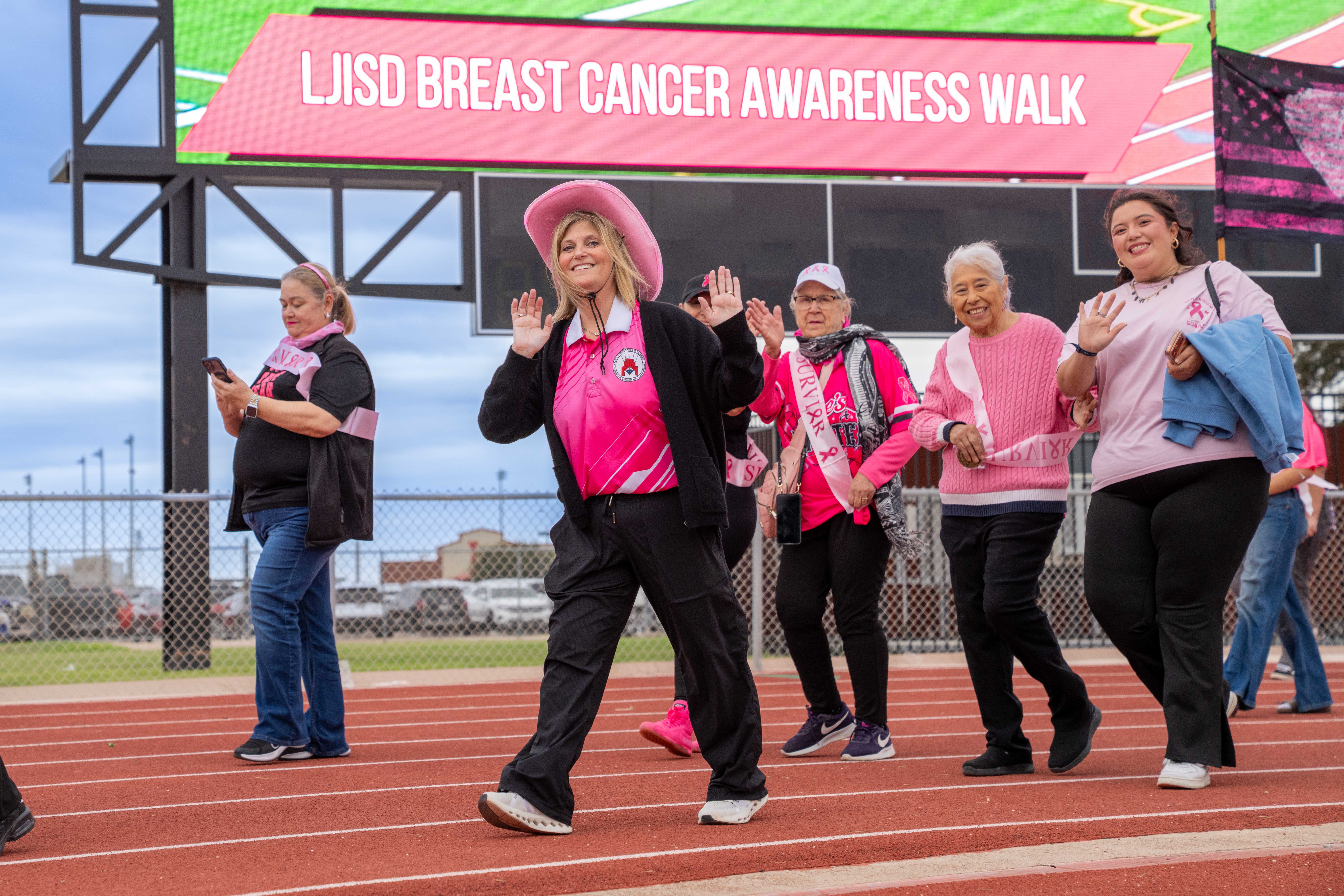 A group of women walk on a track during the "LJISD BREAST CANCER AWARENESS WALK," waving and smiling.