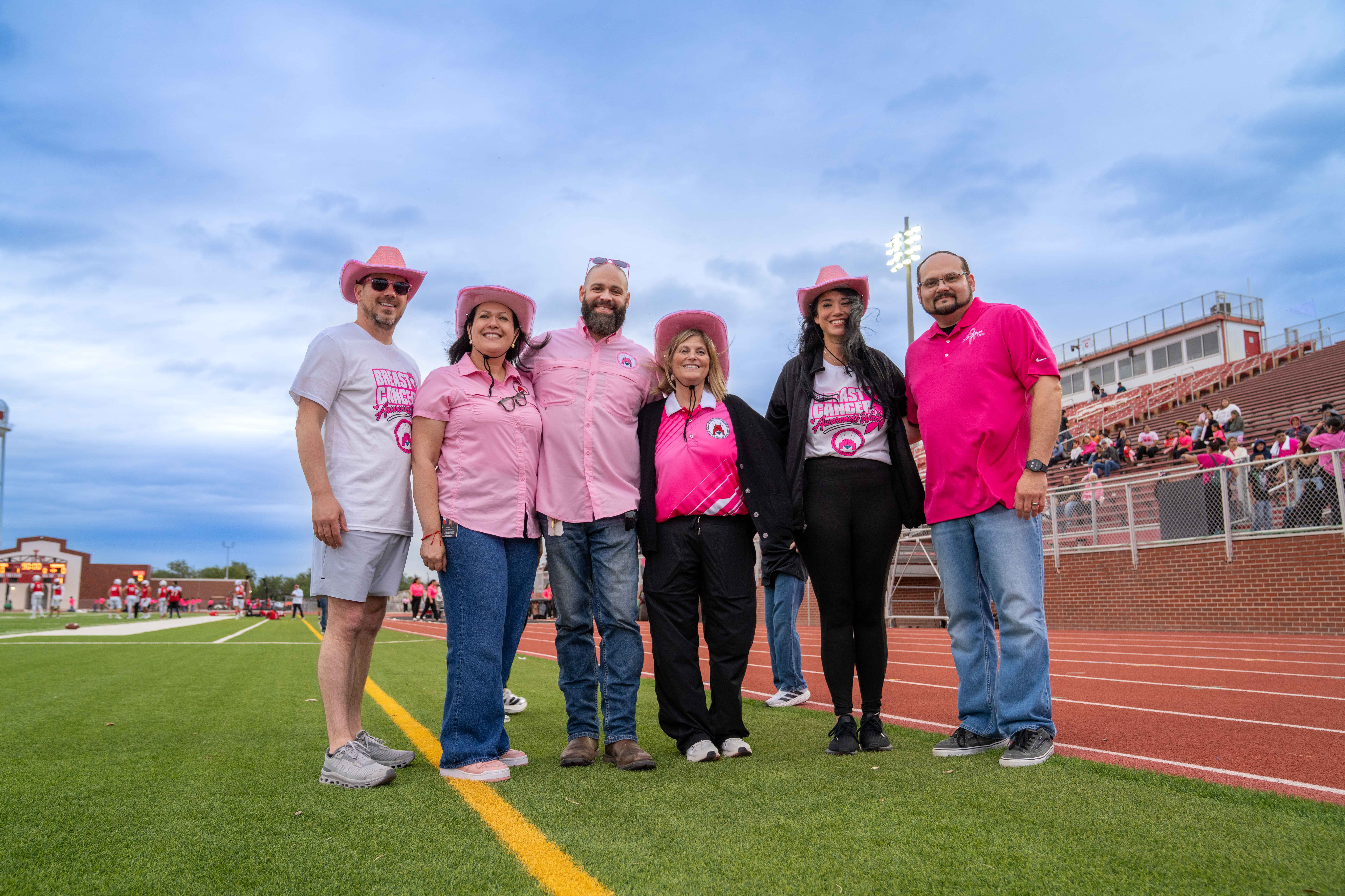 Six adults stand together on a sports field wearing pink clothing and hats to show support for breast cancer awareness.