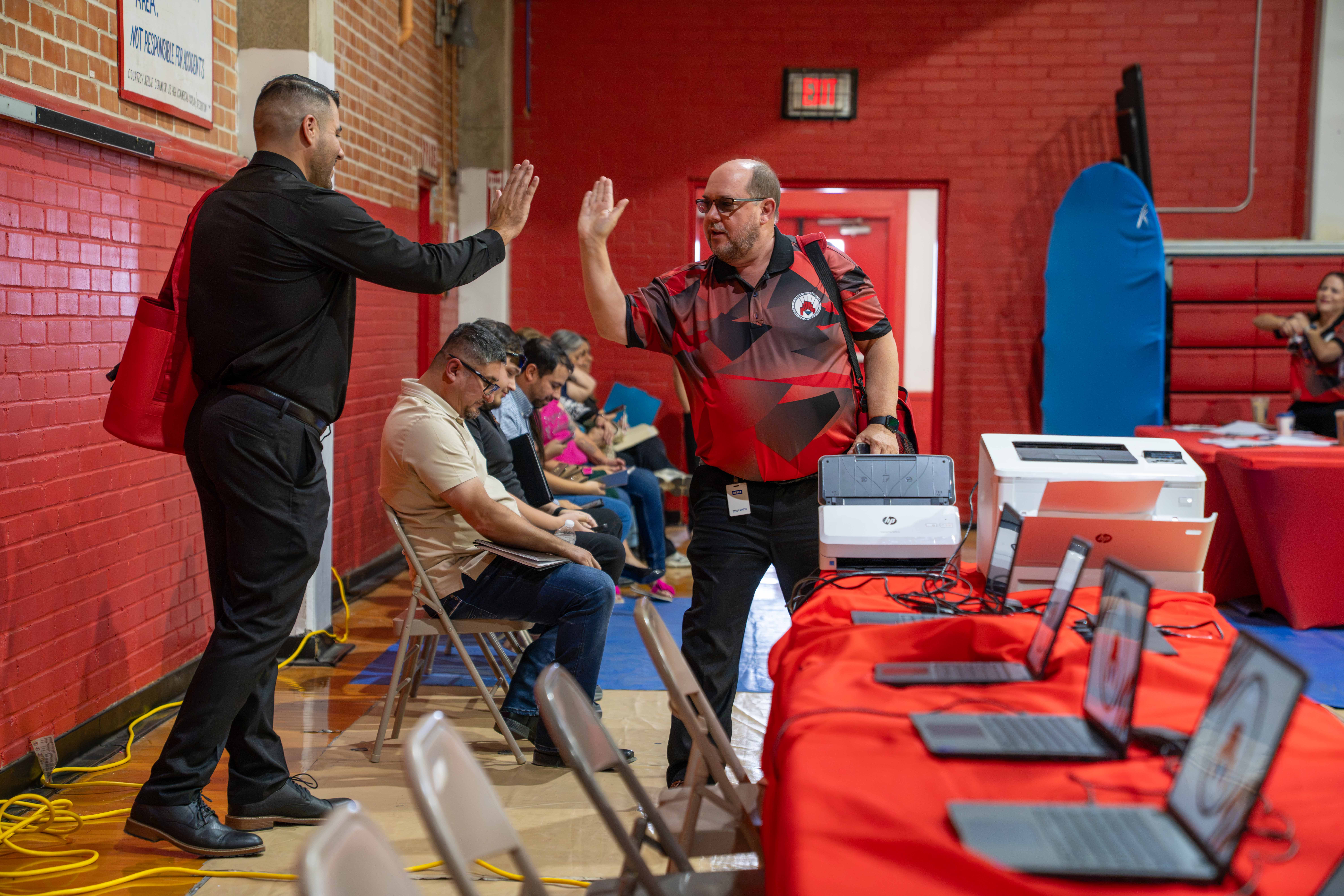 Two men in a gymnasium happily exchange a high-five near a row of tables set up with laptops.