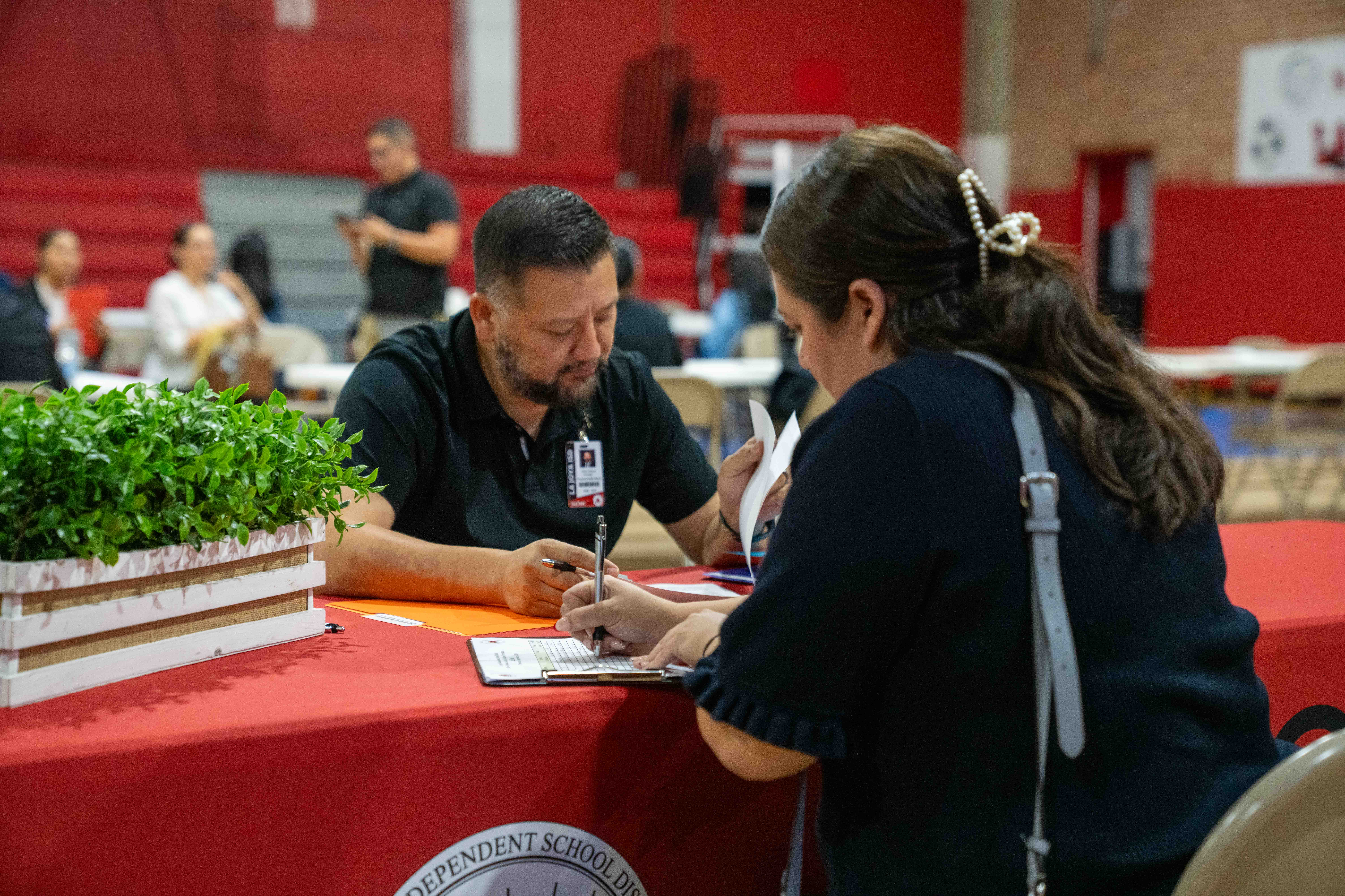 A man and woman review paperwork together at a table covered in a red cloth in a gymnasium.