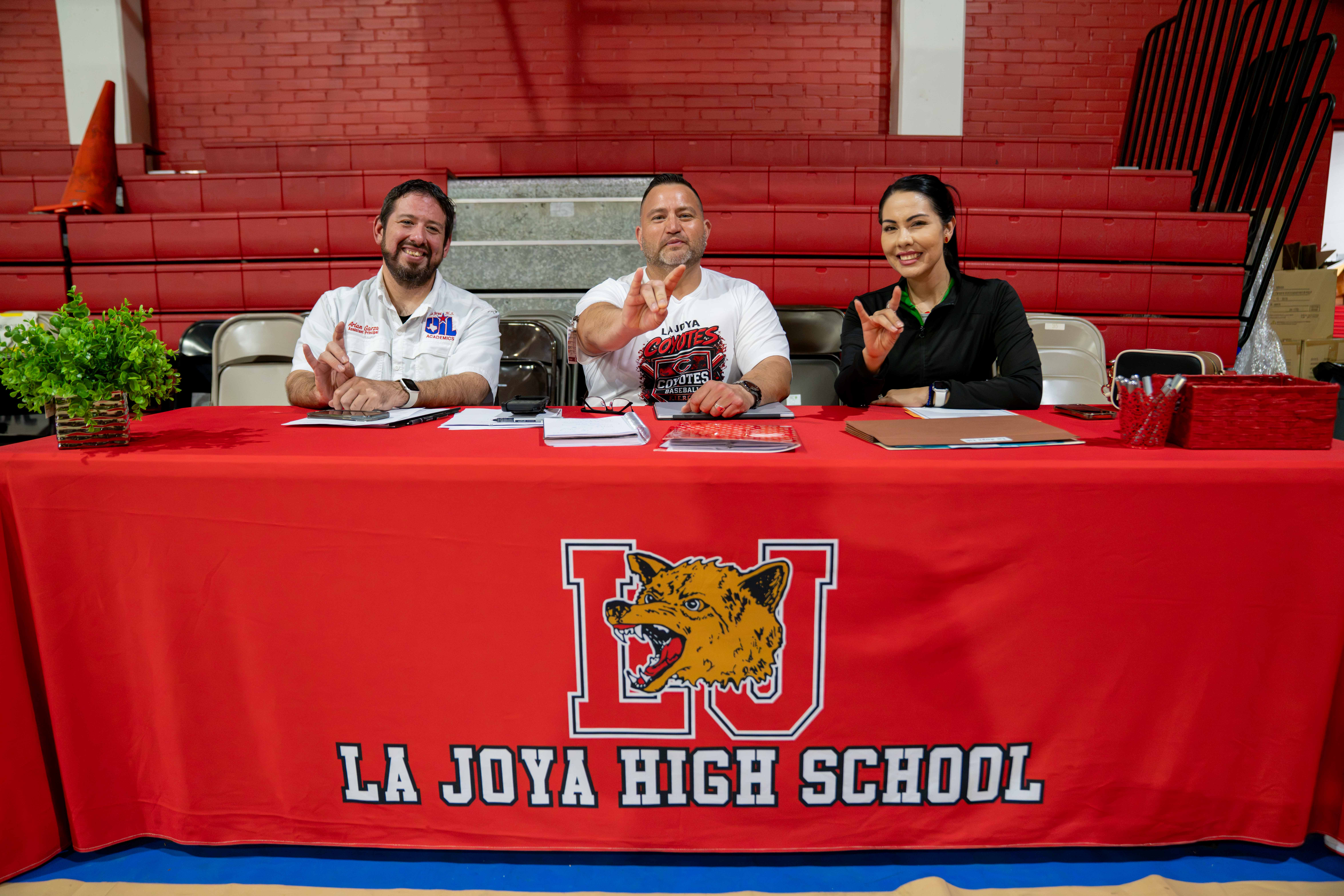 Three people sit behind a red table with the La Joya High School coyote logo, smiling and making hand gestures in a gymnasium.