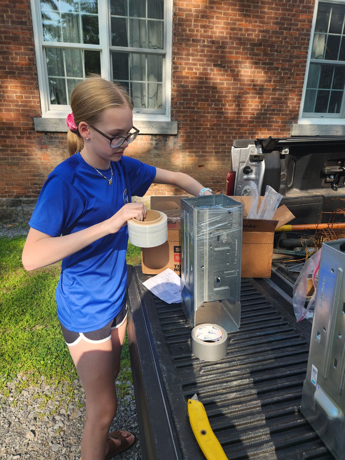 A person wearing glasses and a blue shirt holds a roll of tape next to a truck bed filled with metal HVAC components.