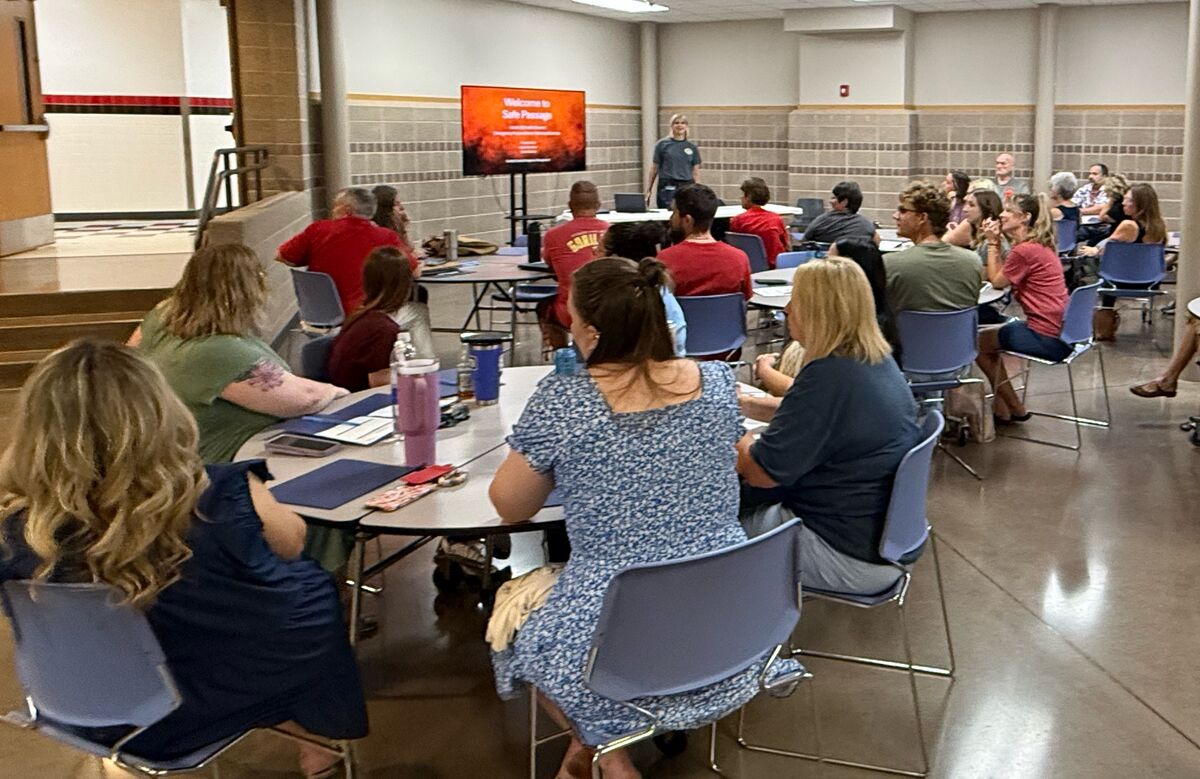 A group of people are seated at tables in a room, listening to a presenter.