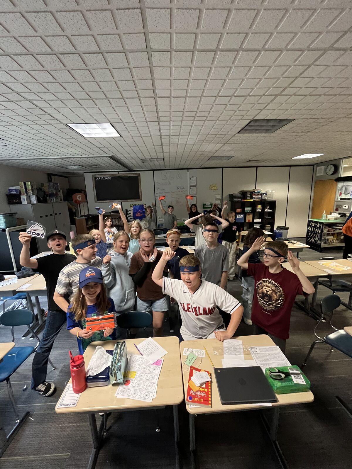 A group of students poses playfully in a brightly lit classroom, holding up various objects and smiling for the camera.