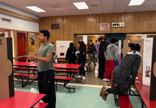 Students present projects at tables set up in a school cafeteria with wood-paneled walls.