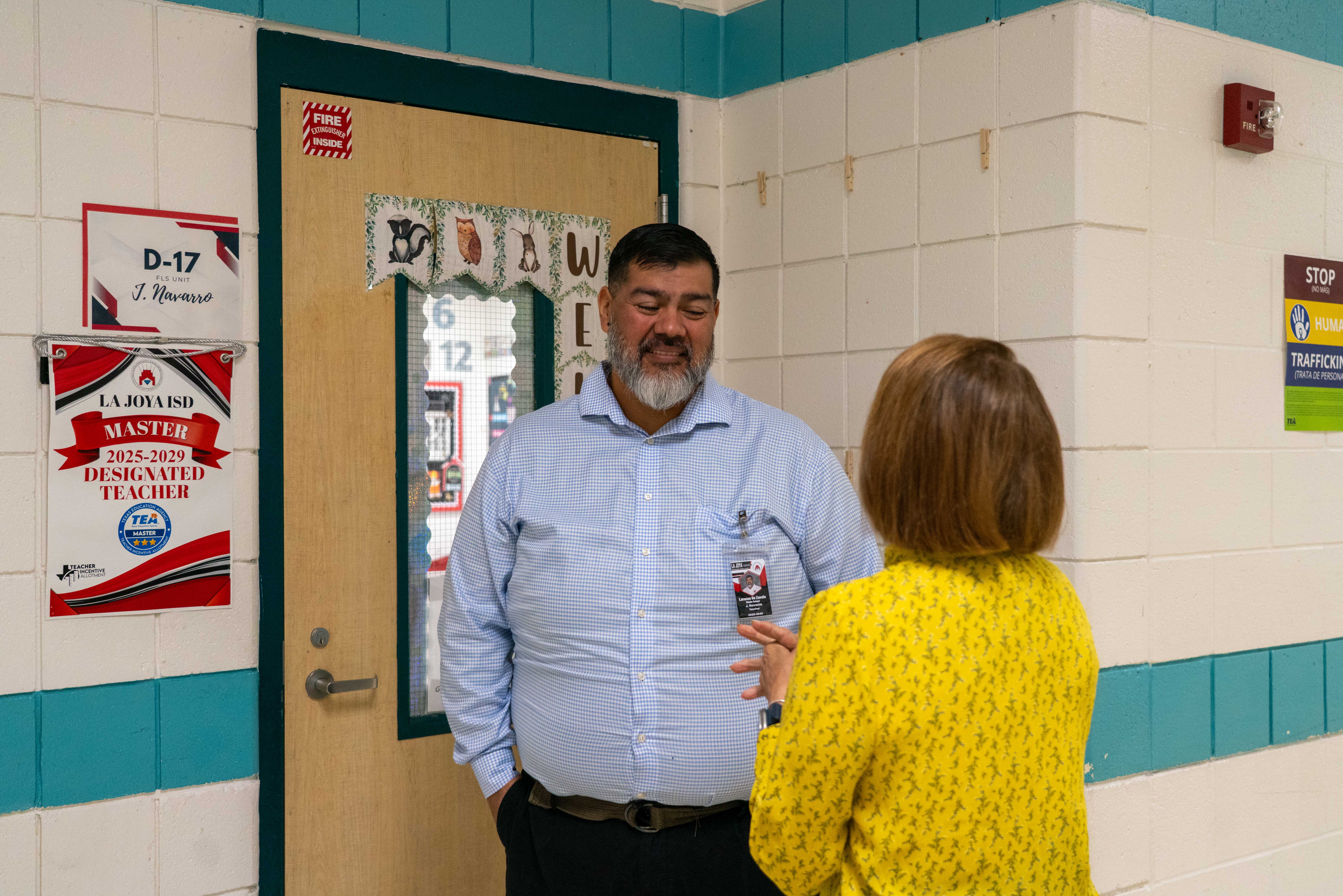 A man in a blue checkered shirt and a woman in a yellow patterned shirt converse in a school hallway.