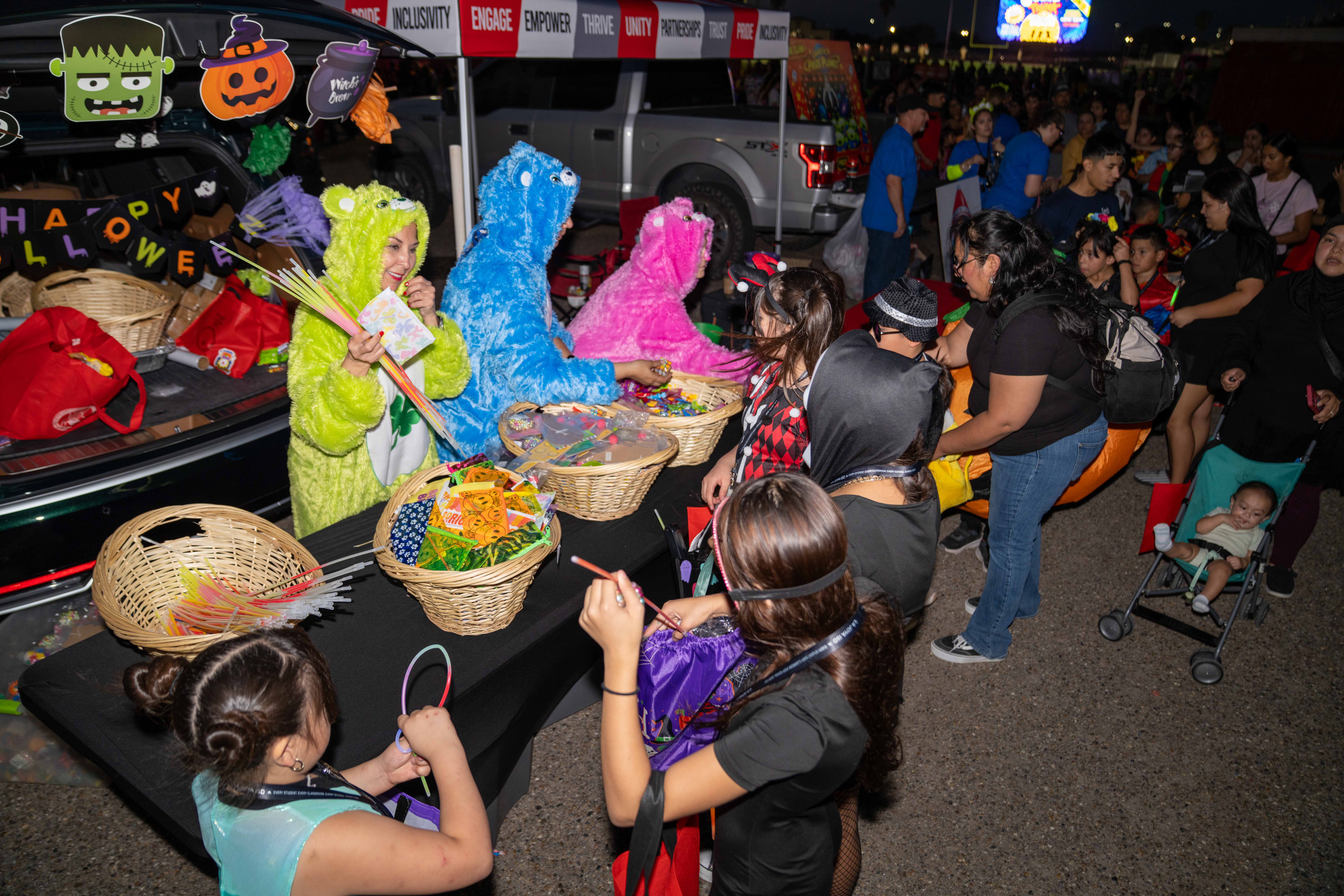 People in costumes hand out treats from a decorated car trunk at an evening Halloween trunk-or-treat event.