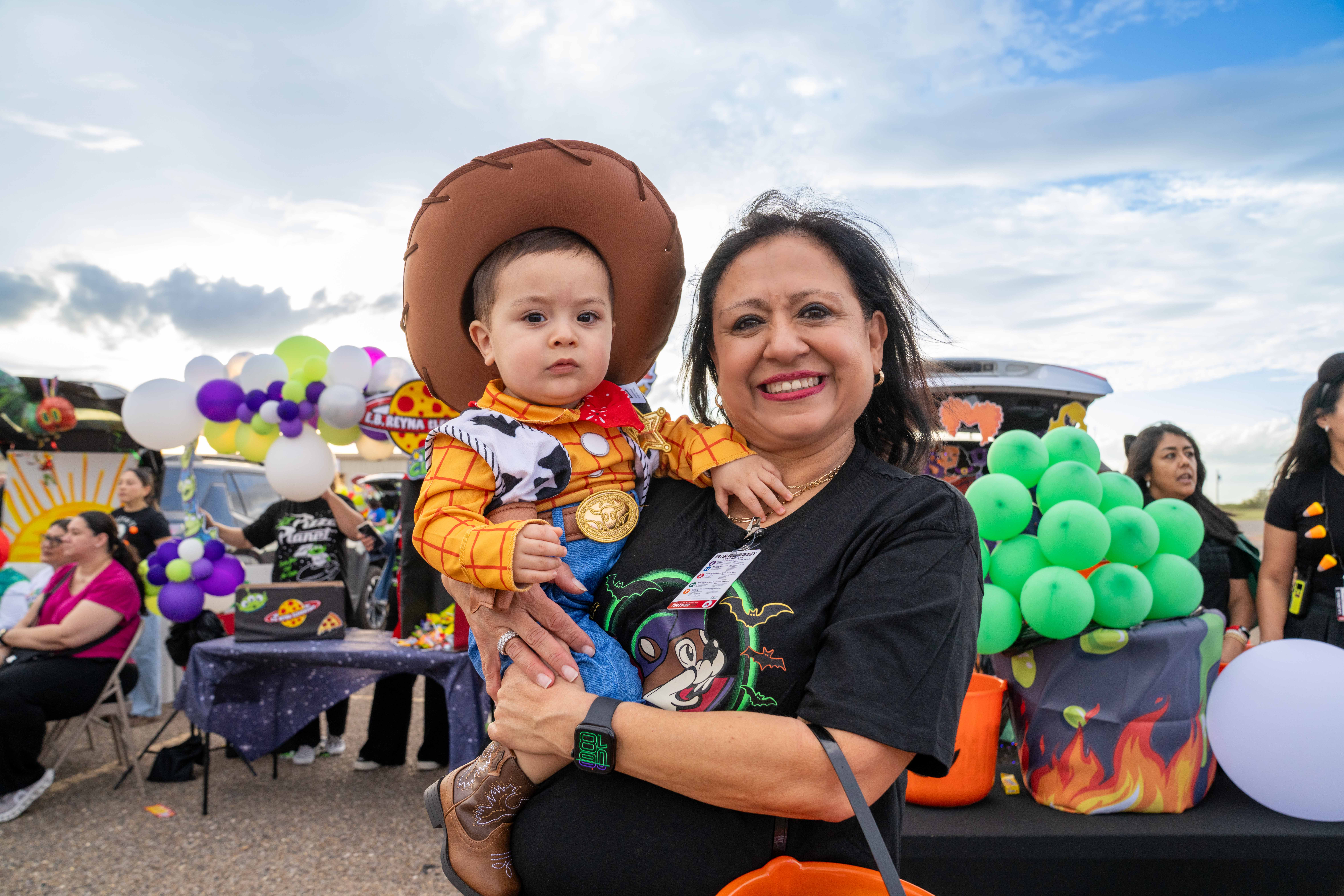 A smiling woman holds a baby dressed as Woody from Toy Story outdoors.