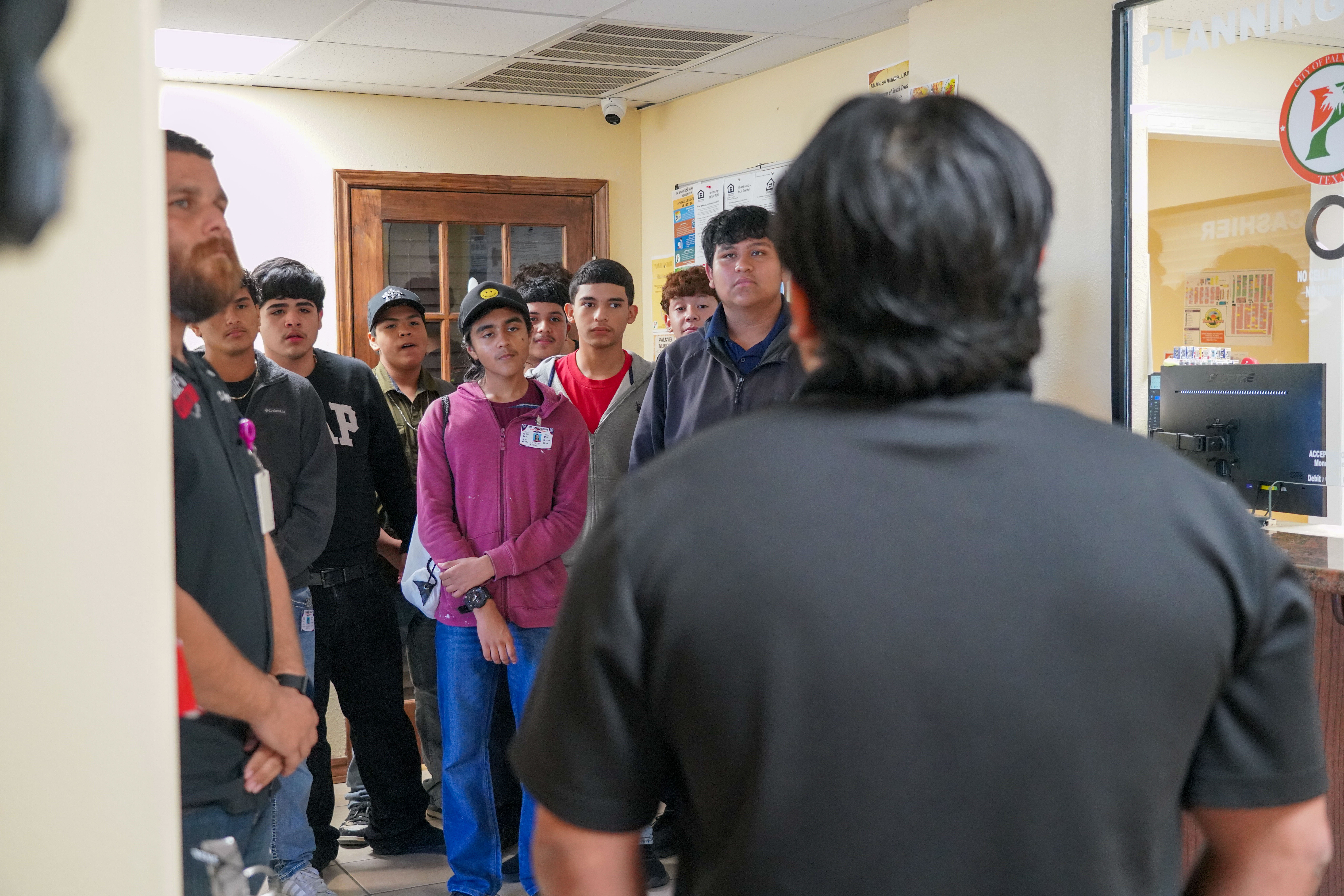A group of young men and a man with a beard stand in a room, listening to someone speak.