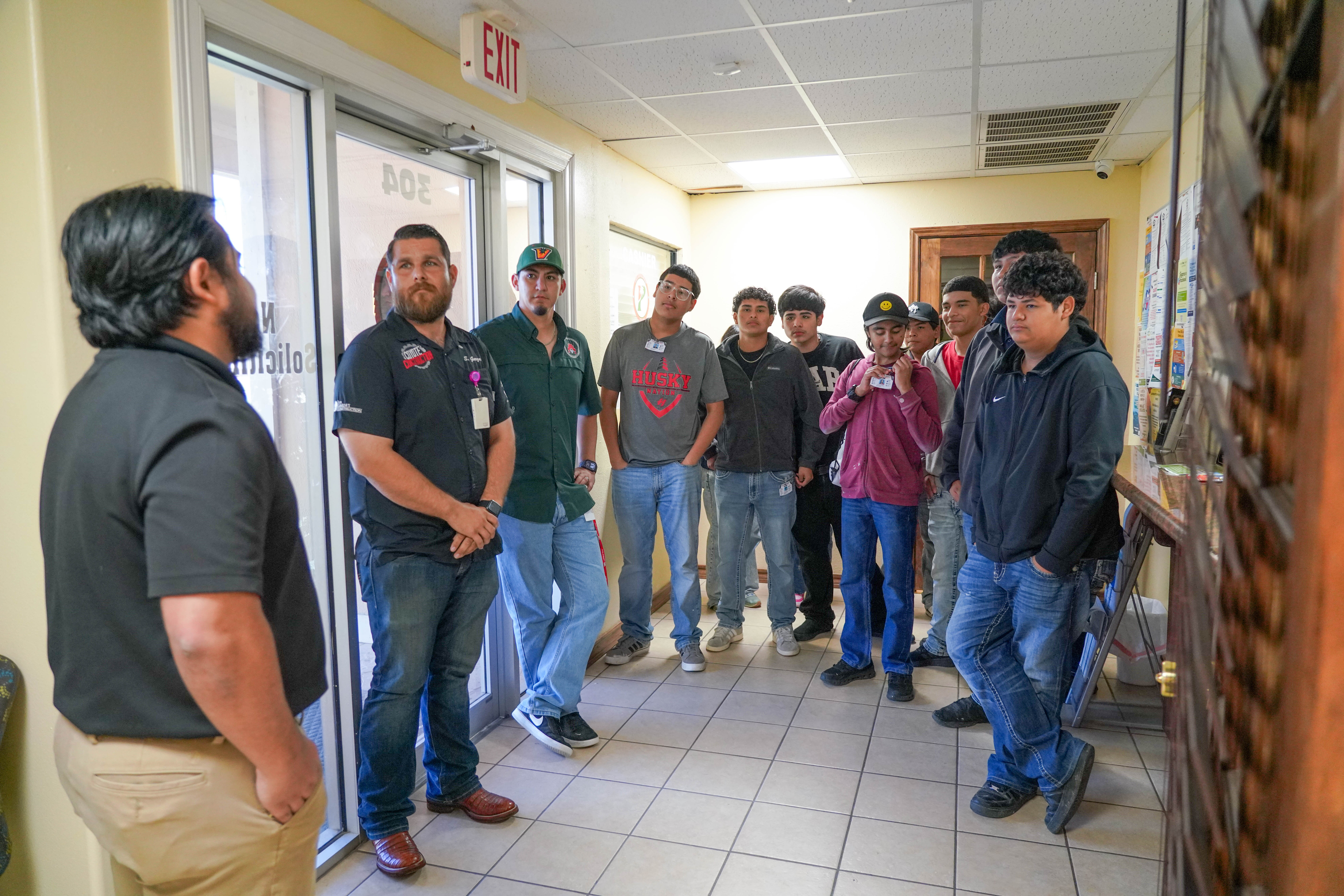 A group of young men and two adults stand in a hallway, appearing to listen to someone speaking.