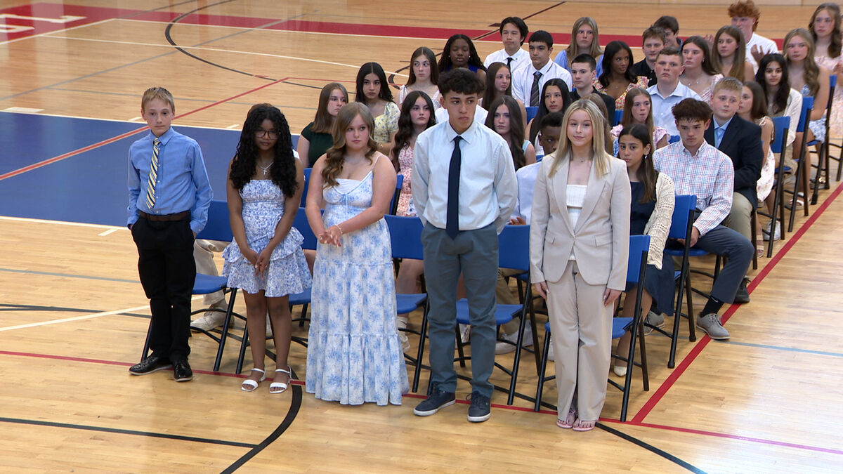 A group of young people in formal attire stand and sit in a gymnasium, facing forward.