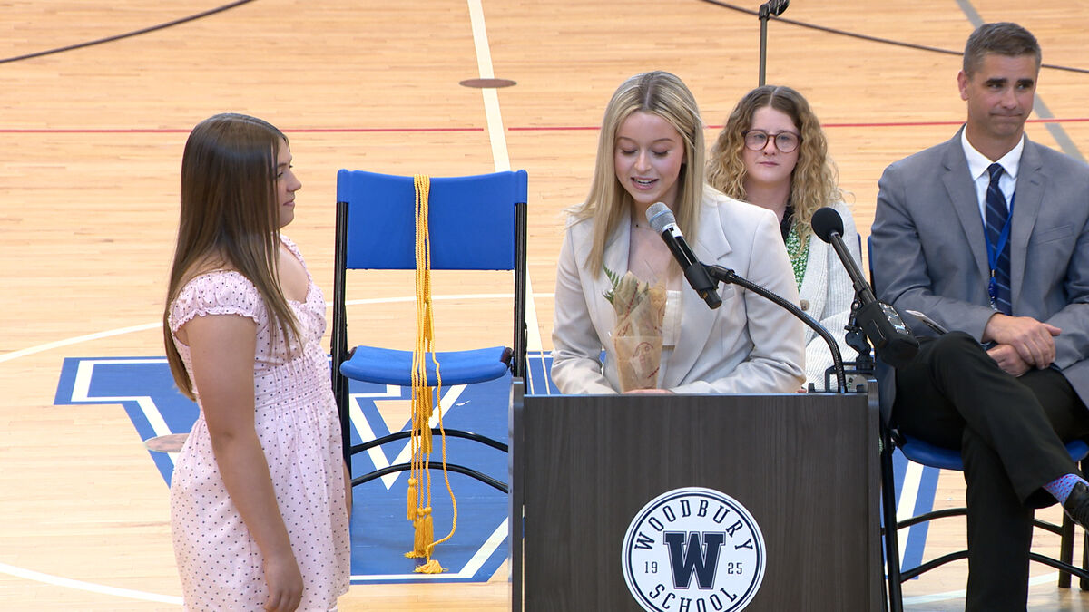 A young woman speaks at a podium with a bouquet of dried flowers, while another young woman stands to her left.