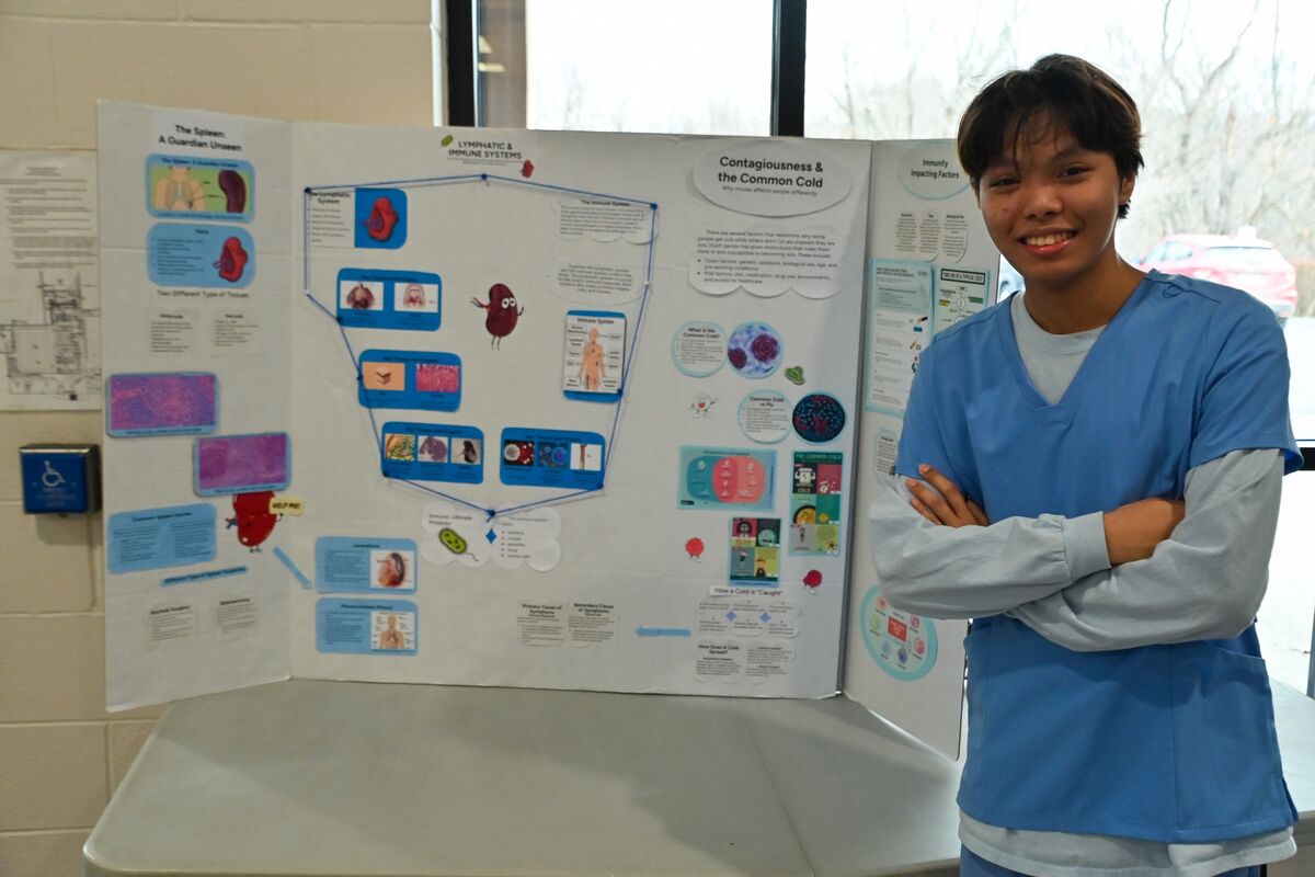 A young person in blue scrubs stands proudly next to a science fair project about the lymphatic and immune systems.