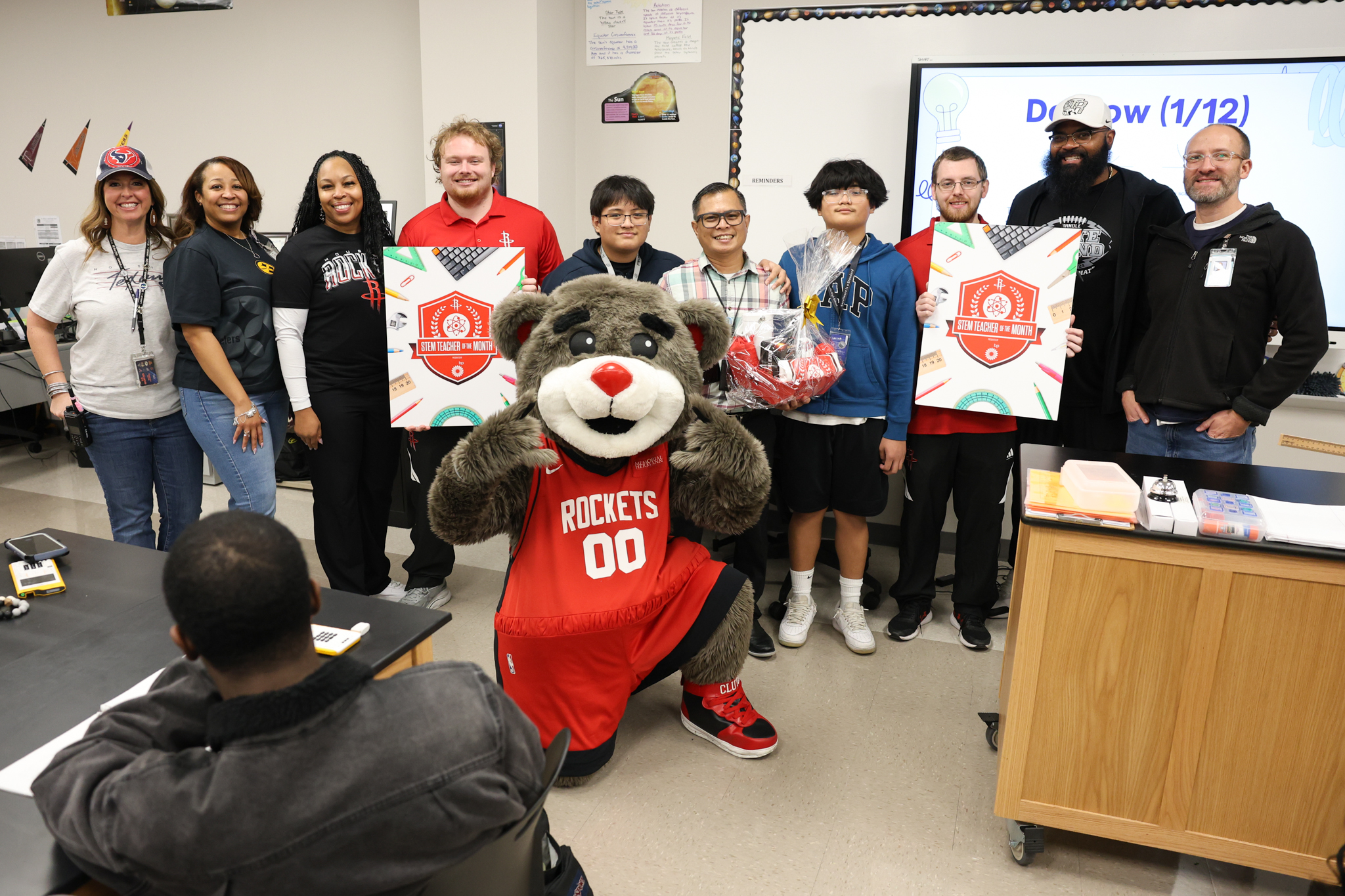 A group of people, including a mascot in a Rockets uniform, pose for a photo in a classroom with STEM Teacher of the Month signs.