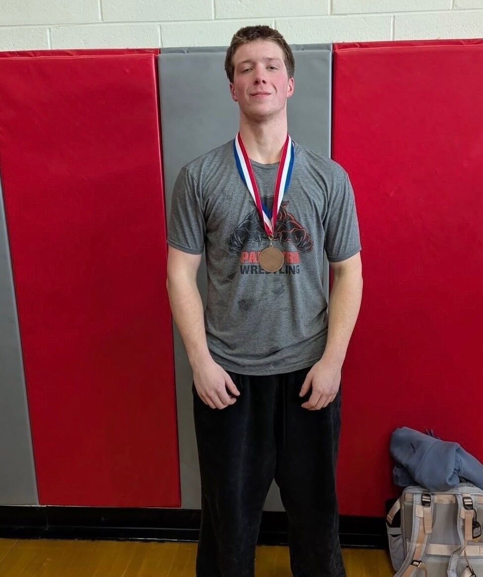 A young man- Senior Wrestler Deakon Haines in a grey wrestling t-shirt and black pants stands proudly with a medal around his neck.