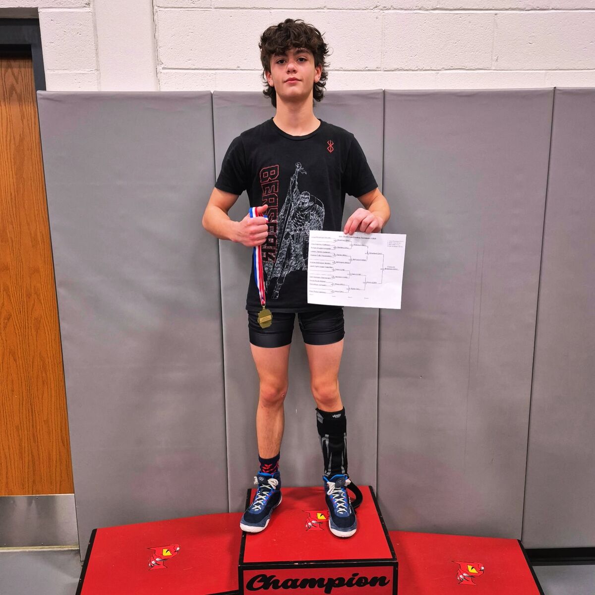 A young wrestler stands on a red podium, holding a medal and a tournament bracket.