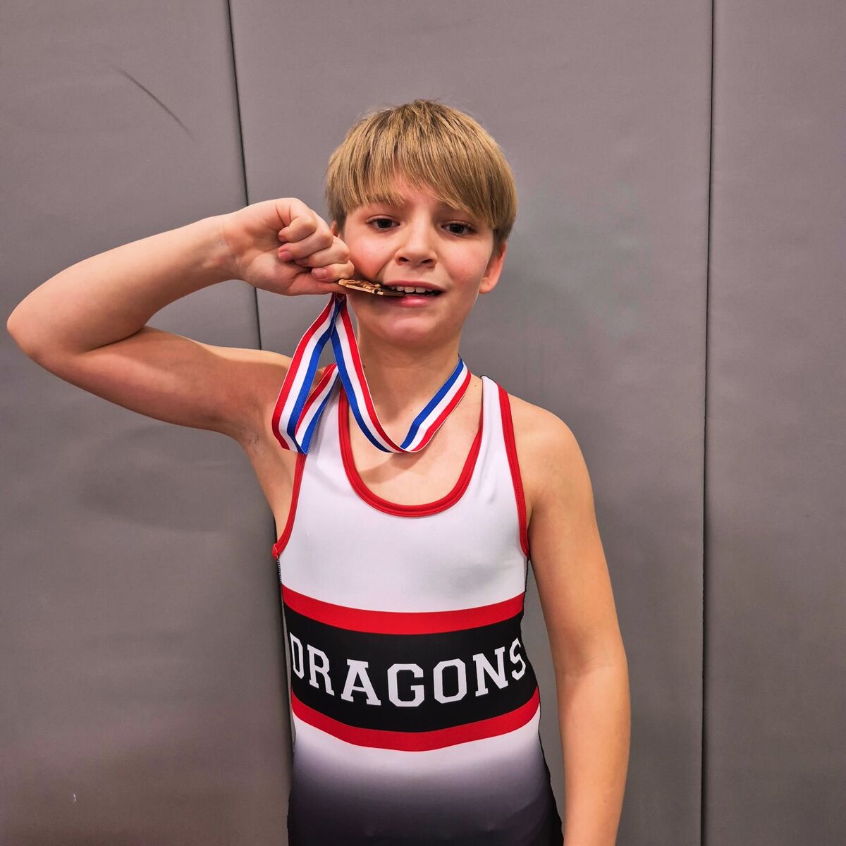 A young wrestler proudly bites a bronze medal with a red, white, and blue ribbon.