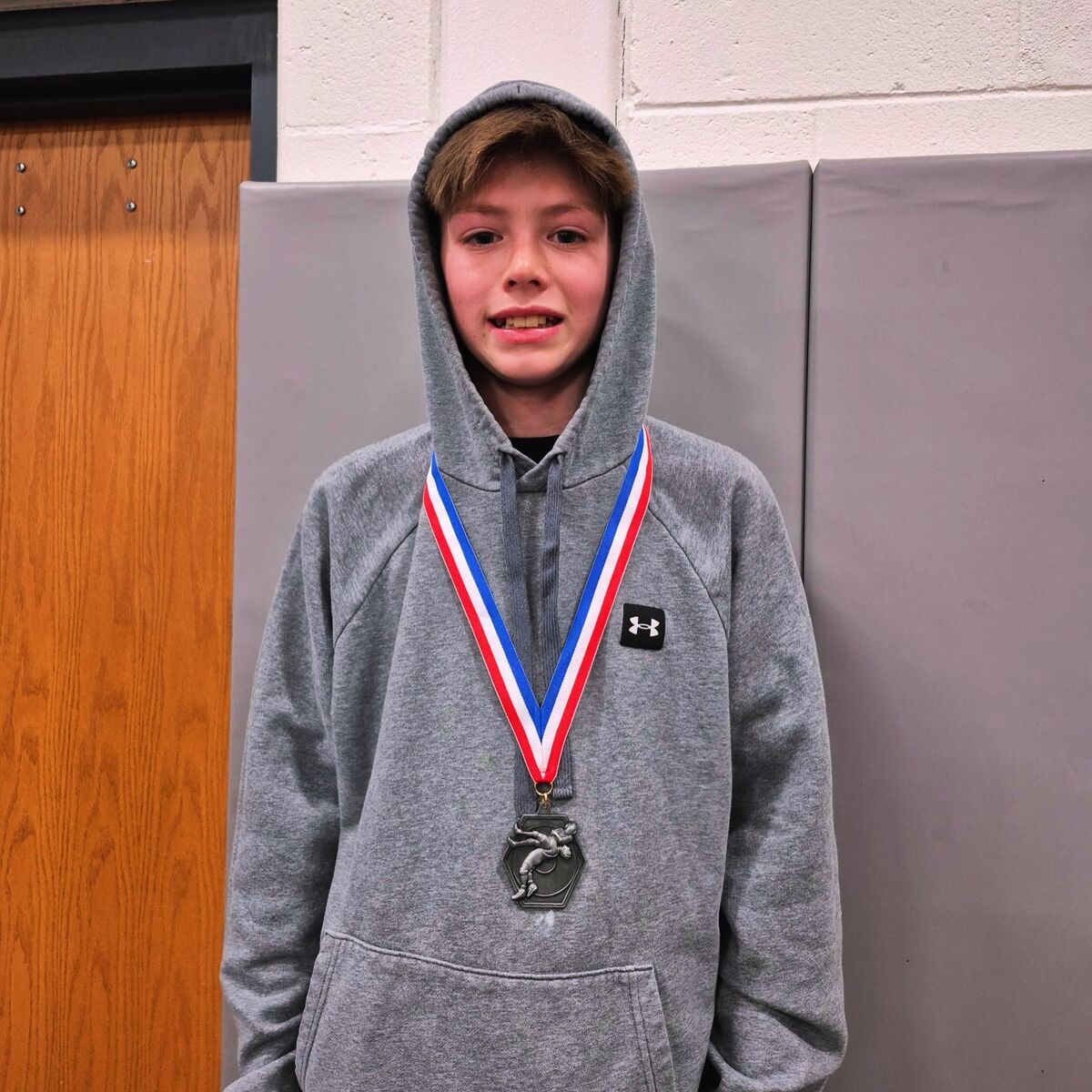 A young person wearing a grey hoodie and a medal stands in front of a padded wall.