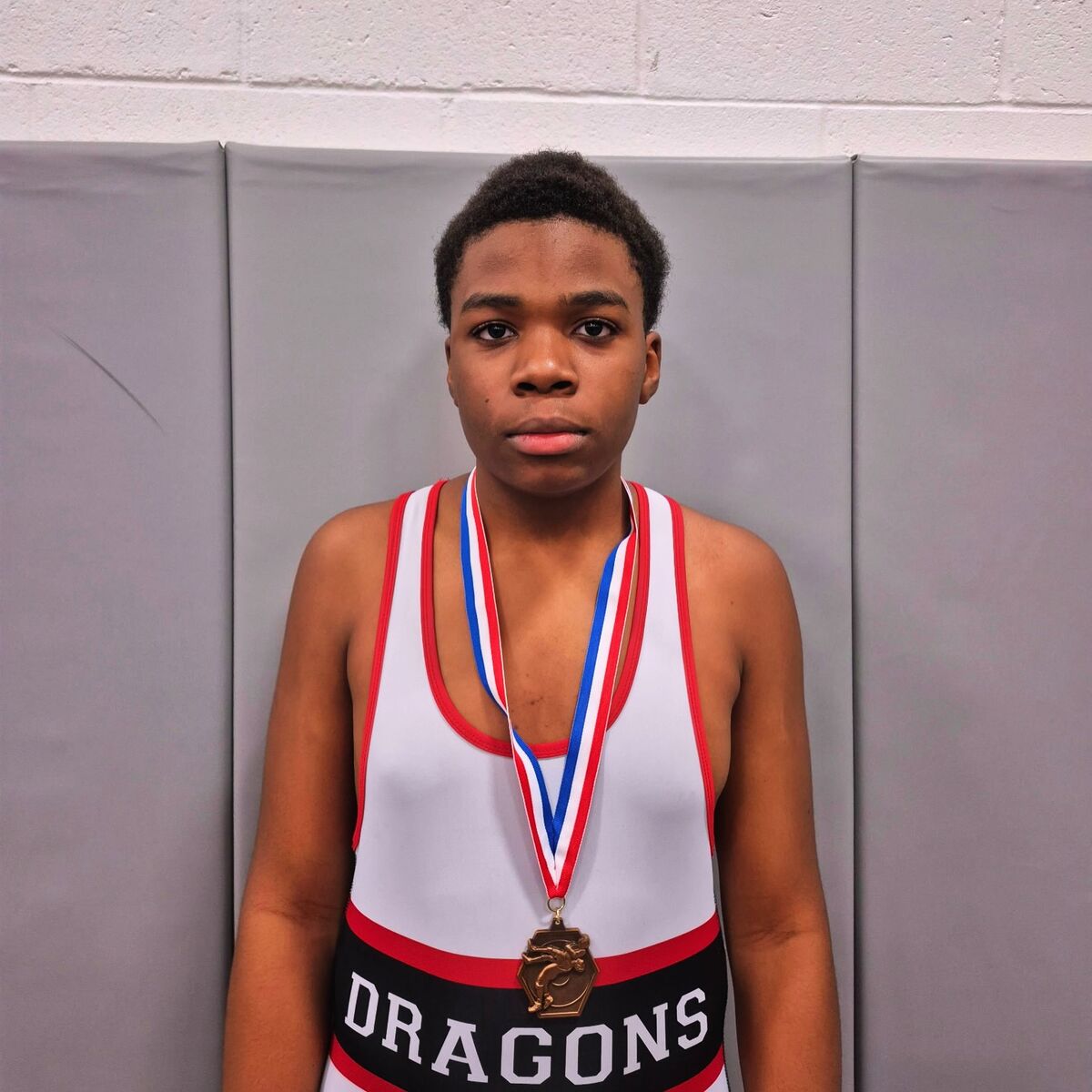 A young wrestler stands proudly wearing a white and red singlet with "DRAGONS" printed on it, and a medal around their neck.