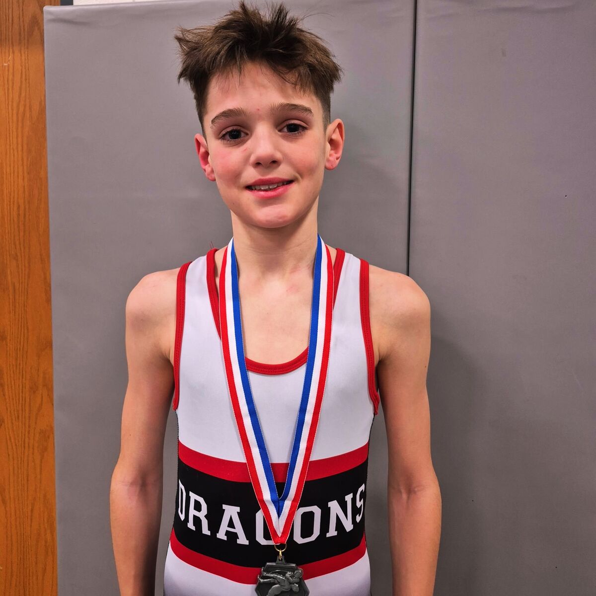 A young wrestler in a white, red, and black singlet with a medal around his neck smiles at the camera.