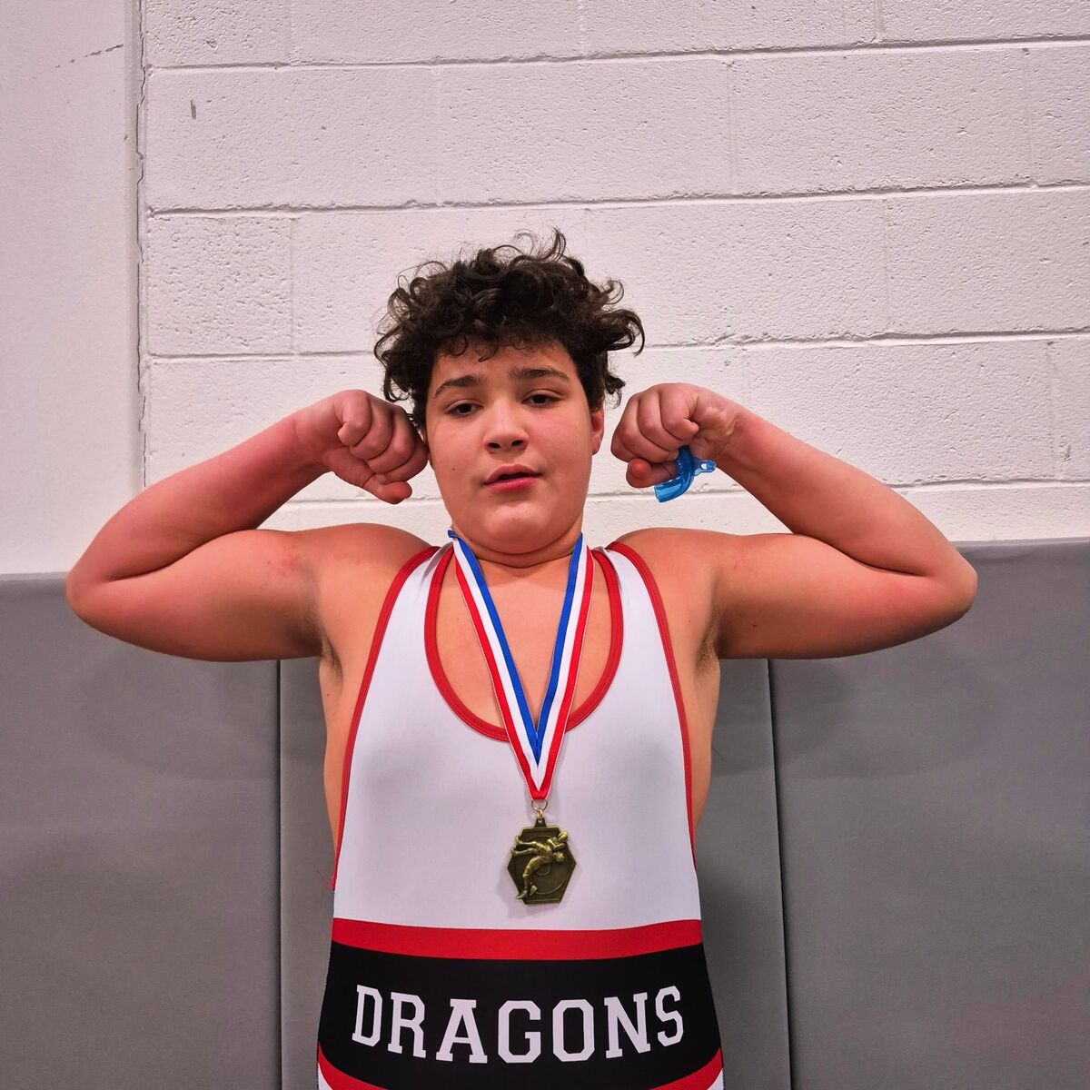 A young wrestler in a white and red singlet with 'DRAGONS' written on it flexes his biceps, wearing a medal around his neck.