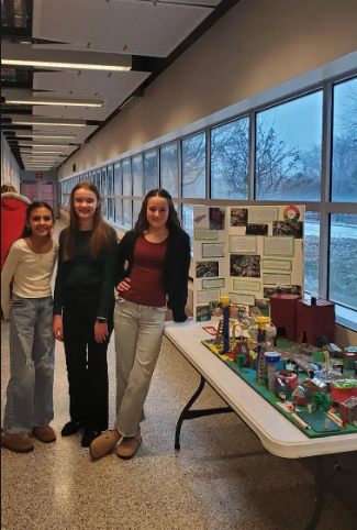 Three young students stand proudly next to a city project display and a presentation board.