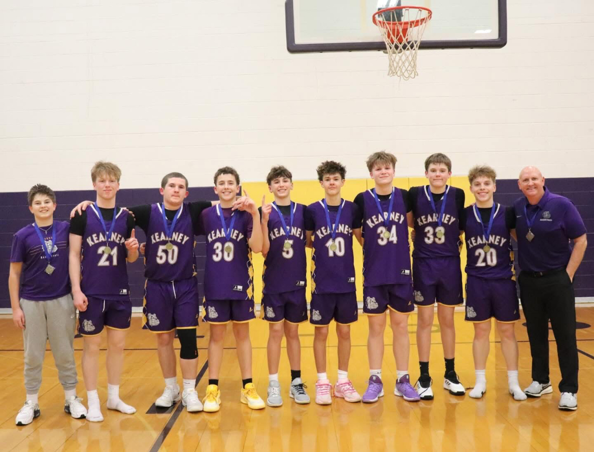 A group of young basketball players in purple jerseys and a coach stand together, all wearing medals.