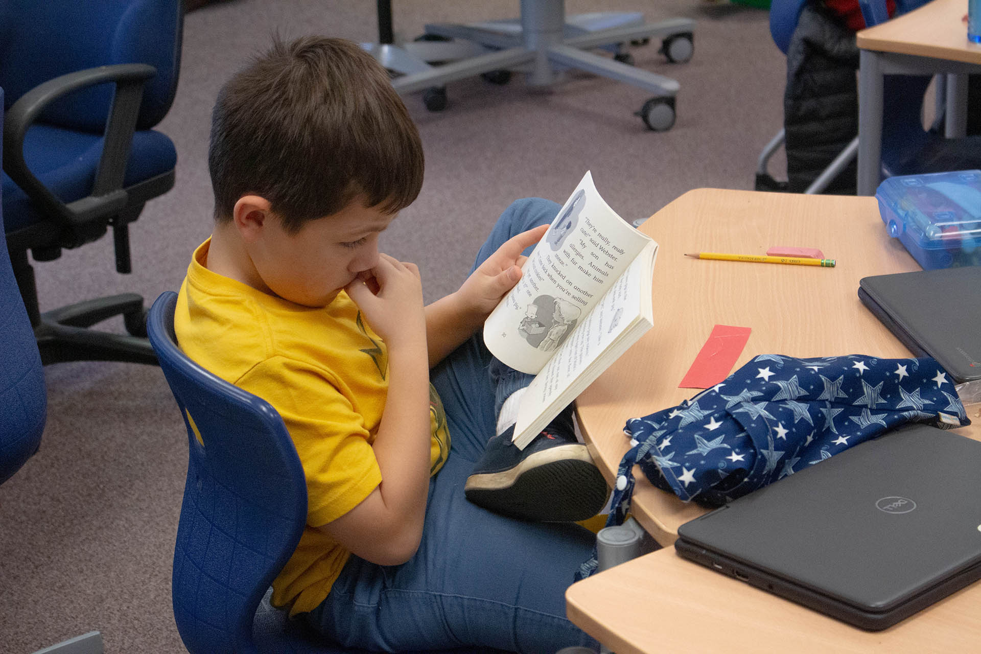 A young boy in a yellow shirt sits at a desk, engrossed in reading a book.