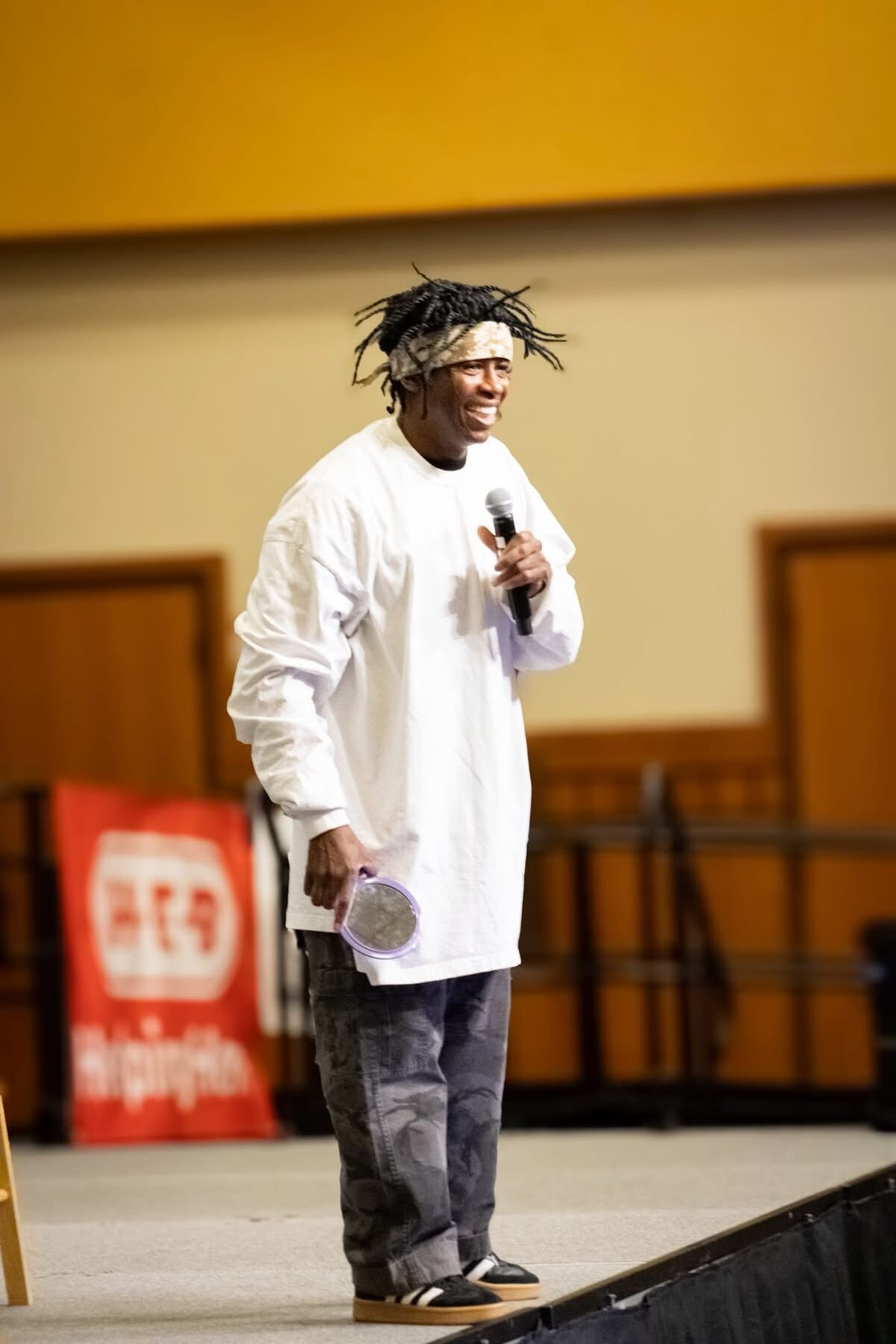 A man with dreadlocks and a bandana smiles while holding a microphone and a small mirror.