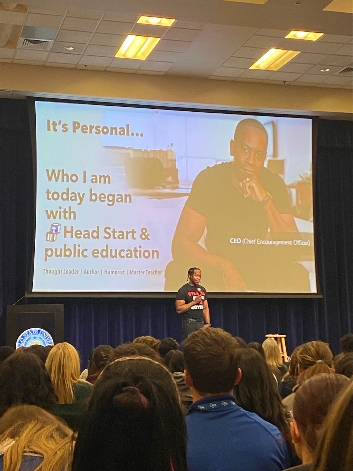 A speaker stands on stage addressing an audience, with a large screen behind him displaying "It's Personal... Who I am today began with Head Start & public education."