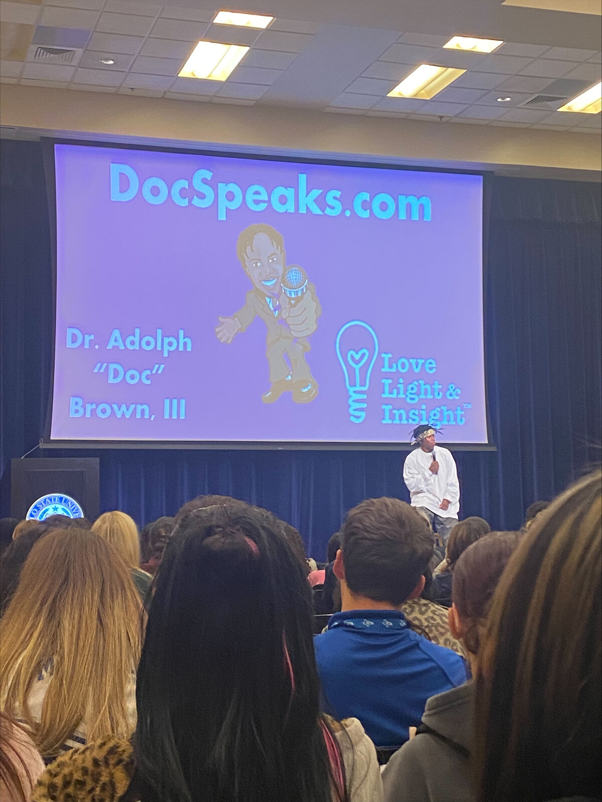 A speaker, Dr. Adolph "Doc" Brown, III, addresses an audience in front of a screen displaying DocSpeaks.com.