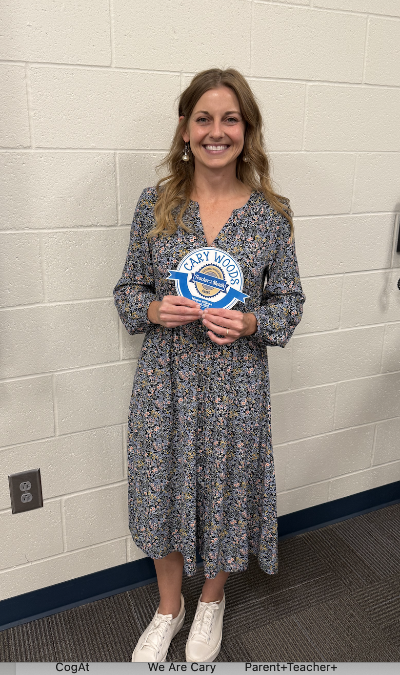 A smiling woman in a floral dress holds a "Cary Woods Teacher of the Month" award.