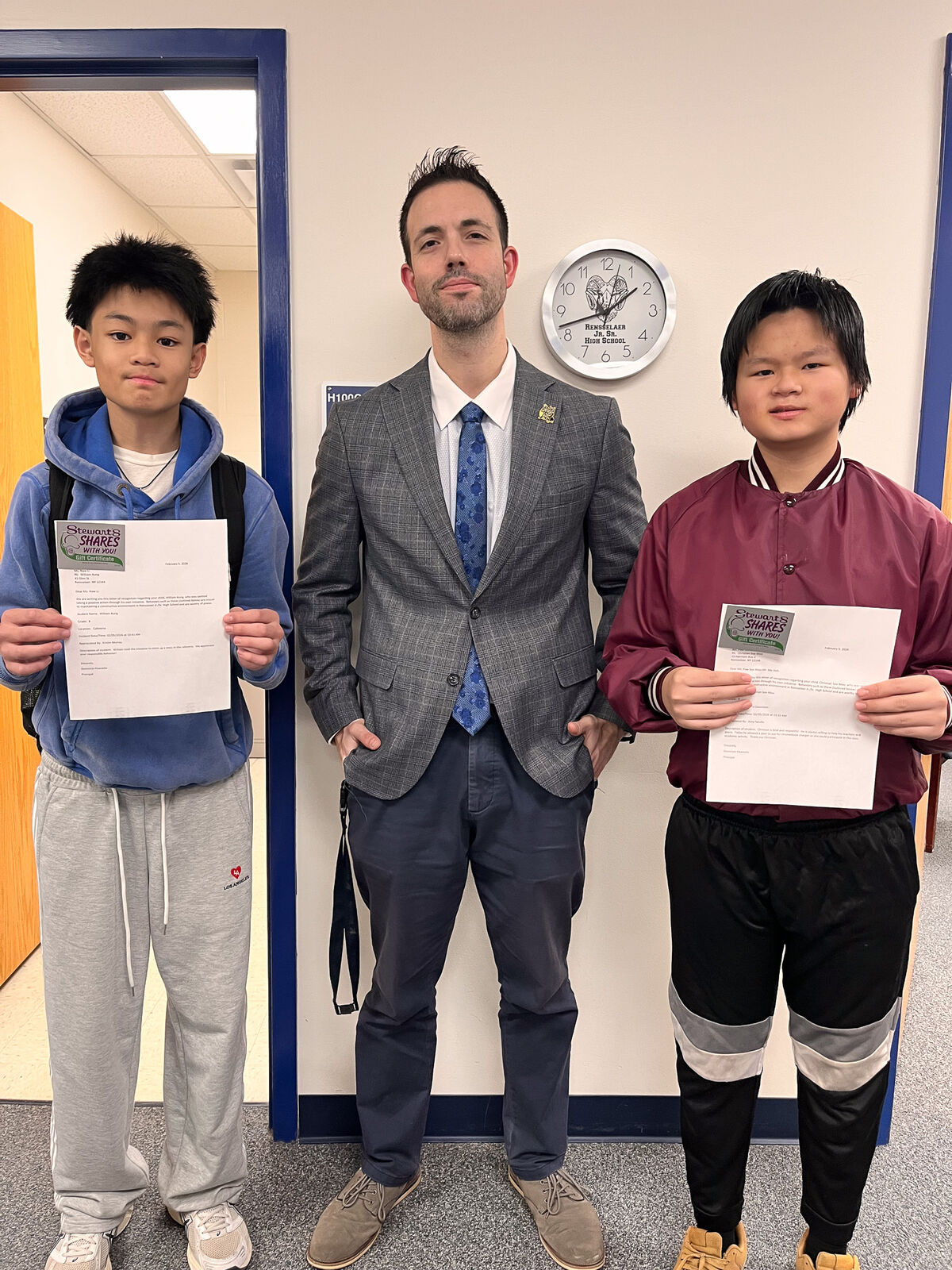 A teacher stands between two students, each holding a certificate from "Stewart's Shares With You!"