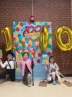 Three children dressed as 100-year-olds celebrate the 100th day of school with festive decorations.
