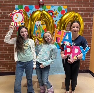 Three smiling girls pose for a photo in front of a '100 Days' balloon display, holding colorful signs.