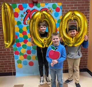 Three smiling boys hold large gold balloons shaped like the number 100 in front of a colorful backdrop celebrating 100 days of school.