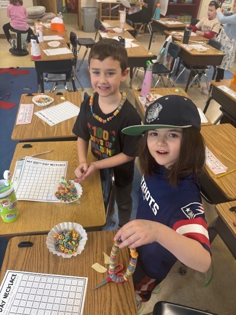 Two children smile while wearing colorful cereal necklaces in a classroom.