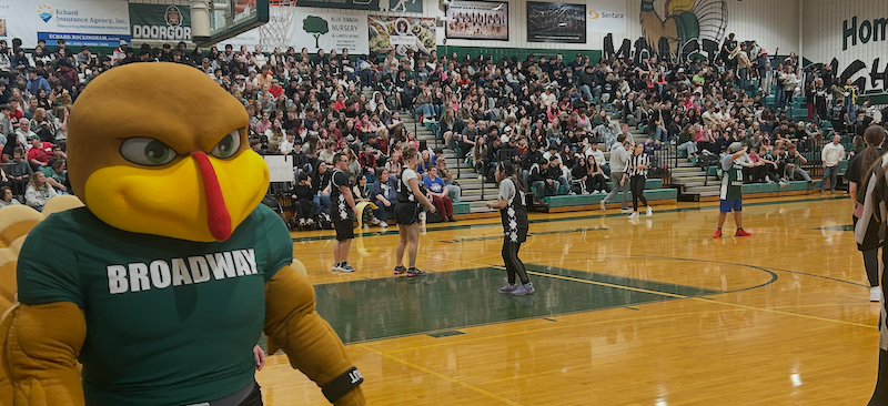 A mascot in a turkey costume with 'BROADWAY' on its shirt stands on a basketball court.