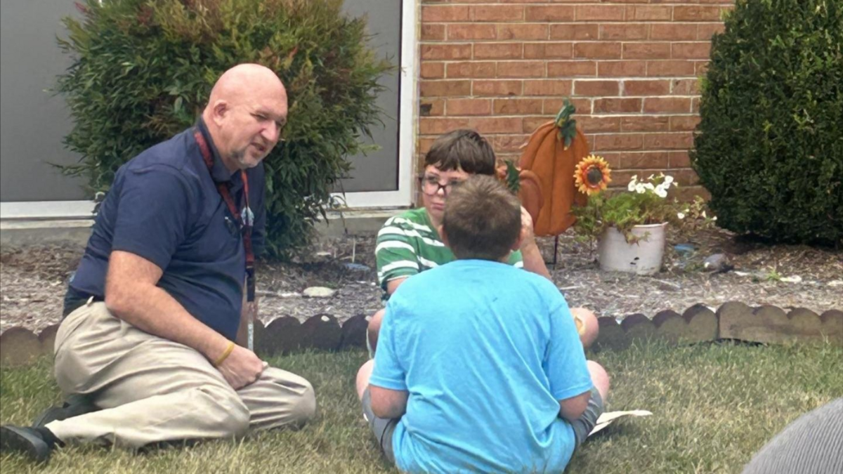 A man in a blue shirt and khaki pants talks to two children sitting on the grass.