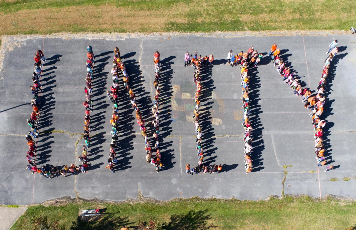 An aerial view shows a group of people forming the word 'UNITY' with their bodies on an asphalt surface.