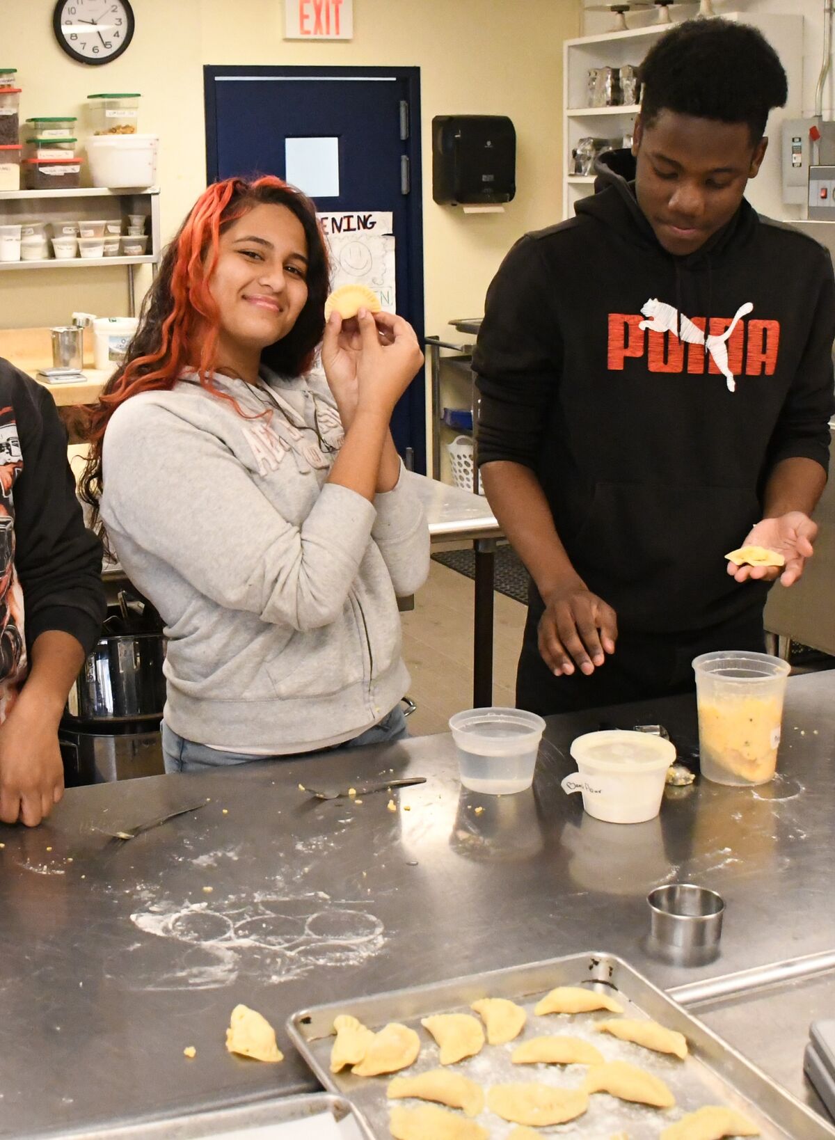 Student Cianna Waiters holds up a pierogi she made as another student makes one in a kitchen classroom.