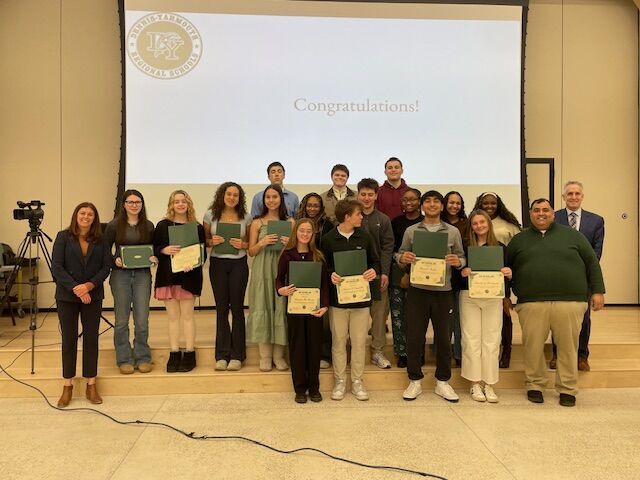 A group of students and adults stand on risers in front of a screen displaying 'Congratulations!' and a school logo.
