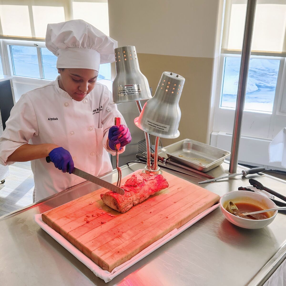 A chef named Alyshah, wearing a white chef's coat and hat, carves a large piece of meat on a wooden cutting board under warming lamps.