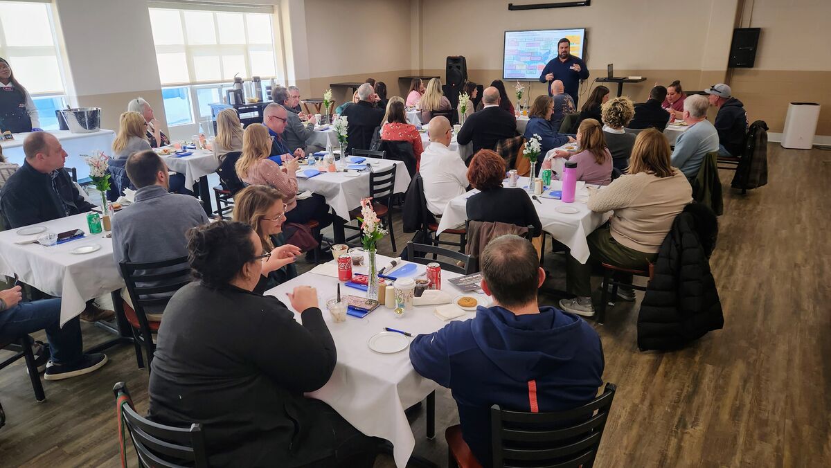 A group of people are seated at tables in a room, listening to a speaker presenting a chart on a screen.