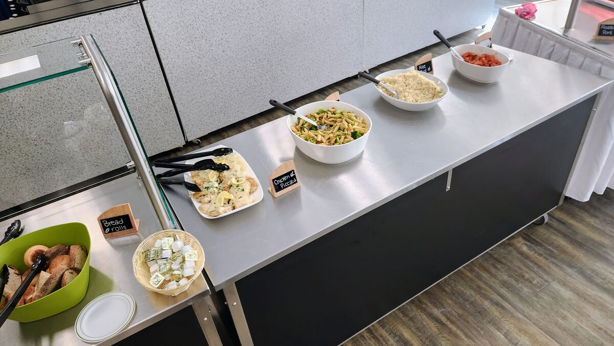 A buffet table displays various dishes including bread rolls, chicken piccata, pasta salad, rice, and chopped tomatoes.
