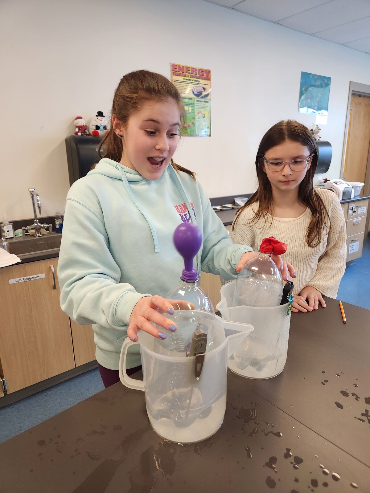 Two girls conduct a science experiment with a bottle, balloon, and measuring cups.