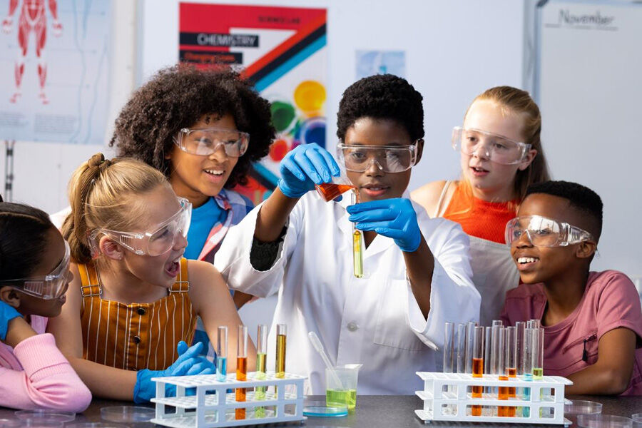 Students in a science lab wear safety goggles as one pours liquid into a test tube.